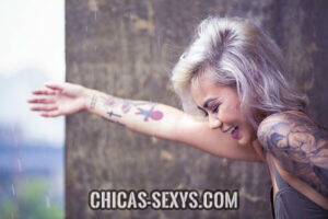 Chicas sexy: Una chica con una sonrisa hermosa posando para una foto. Esta de lado, es decir, solo se ve el perfil de esta mujer joven. Tiene el pelo corto y plateado, tatuajes y una expresión facial atractiva. 